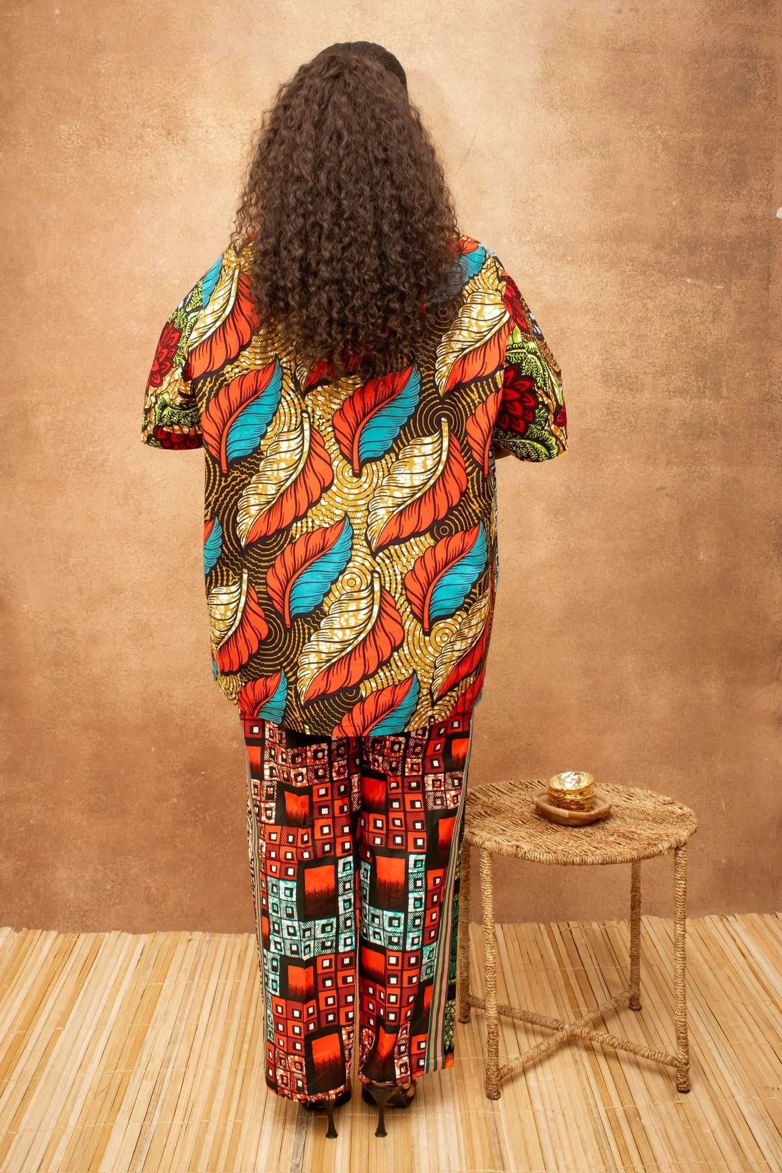 Woman in colourful Ankarza  print outfit, back view, with curly hair and woven stool
